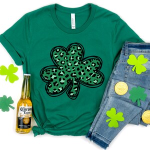 May include: Green t-shirt with a black outline of a shamrock with a green and black leopard print inside. The shirt is laying on a white surface with a bottle of Corona Extra beer and a pair of blue jeans with green shamrock cutouts.