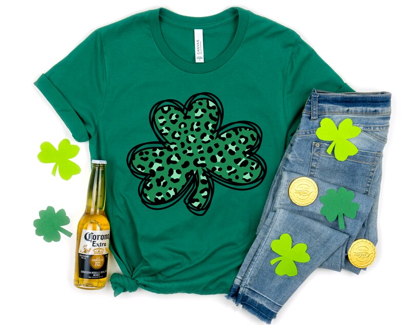 May include: Green t-shirt with a black outline of a shamrock with a green and black leopard print inside. The shirt is laying on a white surface with a bottle of Corona Extra beer and a pair of blue jeans with green shamrock cutouts.