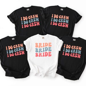 Puede incluir: Cinco camisetas, cuatro negras y una blanca, con texto retro. Las camisetas negras dicen "I DO CREW" en naranja, azul y rosa. La camiseta blanca dice "BRIDE" en los mismos colores. Todas las camisetas tienen mangas cortas.