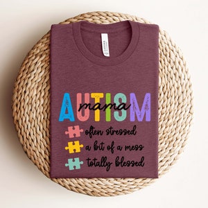 K&ouml;nnte beinhalten: Ein maroonfarbenes T-Shirt mit dem Text "AUTISM mama" in Regenbogenfarben. Dem Text folgen drei Puzzle-Teile mit dem Text "oft gestresst", "ein bisschen ein Chaos" und "total gesegnet".