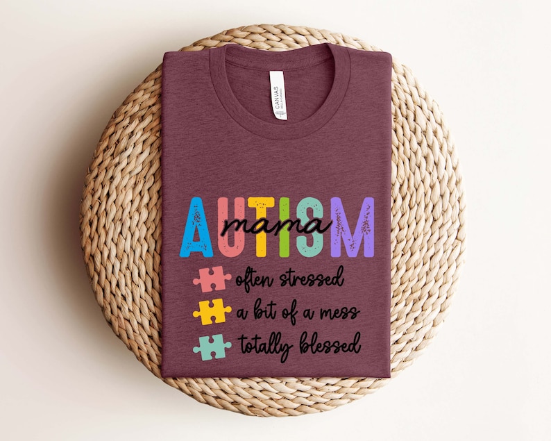 K&ouml;nnte beinhalten: Ein maroonfarbenes T-Shirt mit dem Text "AUTISM mama" in Regenbogenfarben. Dem Text folgen drei Puzzle-Teile mit dem Text "oft gestresst", "ein bisschen ein Chaos" und "total gesegnet".