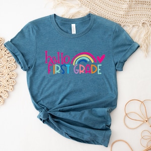 Op de afbeelding: Tealblauw T-shirt met een regenboog en de tekst "hello first grade" in kleurrijke letters.