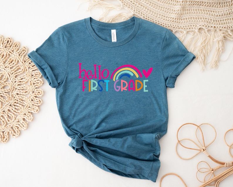 Op de afbeelding: Tealblauw T-shirt met een regenboog en de tekst "hello first grade" in kleurrijke letters.