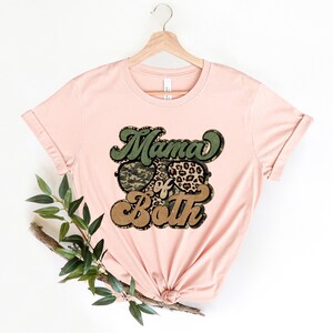 May include: A pink t-shirt with a graphic design that says "Mama of Both" in a retro font. The graphic features a camouflage heart and a leopard print heart.