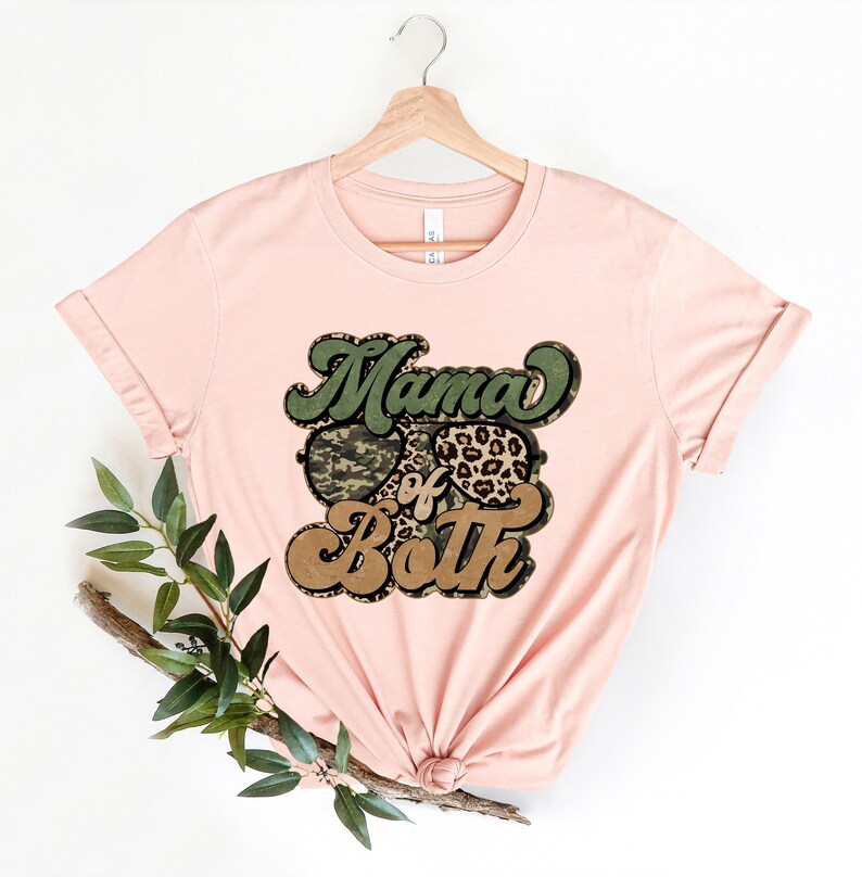May include: A pink t-shirt with a graphic design that says "Mama of Both" in a retro font. The graphic features a camouflage heart and a leopard print heart.