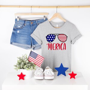 May include: A grey t-shirt with a graphic of sunglasses featuring red, white and blue stars and stripes. The text 'MERICA is printed in red below the sunglasses.