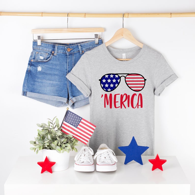 May include: A grey t-shirt with a graphic of sunglasses featuring red, white and blue stars and stripes. The text 'MERICA is printed in red below the sunglasses.