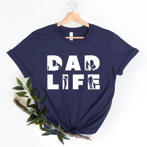 May include: Navy blue t-shirt with white text that reads "DAD LIFE". The letters are made up of silhouettes of a father doing various activities with his child, such as playing ball, pushing a stroller, and holding a child's hand.