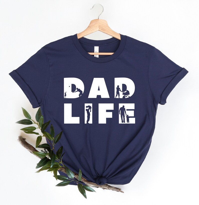 May include: Navy blue t-shirt with white text that reads "DAD LIFE". The letters are made up of silhouettes of a father doing various activities with his child, such as playing ball, pushing a stroller, and holding a child's hand.