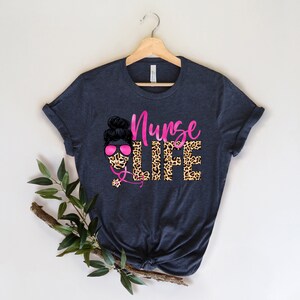 May include: A dark blue t-shirt with a pink and black graphic design. The graphic features a woman with a bun in her hair wearing a leopard print face mask and pink sunglasses. The text "Nurse Life" is written in pink letters with a leopard print background.