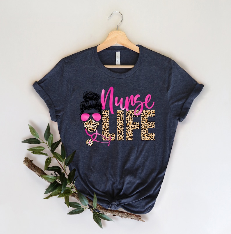 May include: A dark blue t-shirt with a pink and black graphic design. The graphic features a woman with a bun in her hair wearing a leopard print face mask and pink sunglasses. The text "Nurse Life" is written in pink letters with a leopard print background.