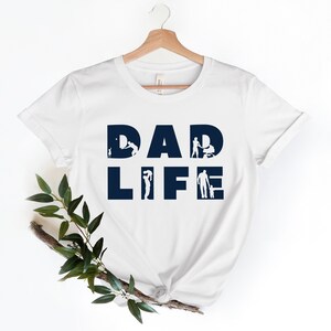 May include: White t-shirt with the text "DAD LIFE" in navy blue. The letters are filled with silhouettes of a father interacting with his children.