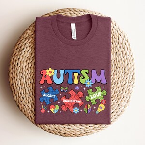 May include: A maroon t-shirt with the word "AUTISM" in colorful letters. The letters are surrounded by puzzle pieces with the words "ACCEPT", "UNDERSTAND", and "LOVE" on them. There are also butterflies, flowers, and other decorative elements.