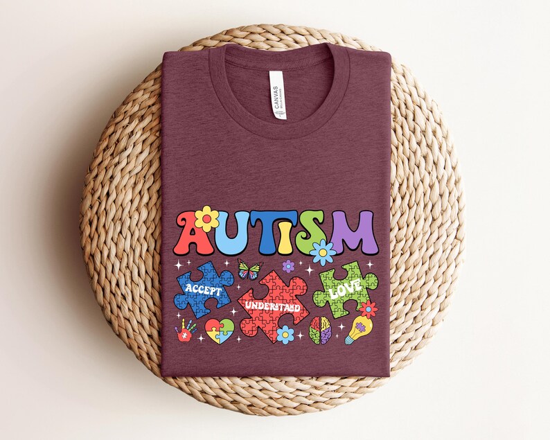 May include: A maroon t-shirt with the word "AUTISM" in colorful letters. The letters are surrounded by puzzle pieces with the words "ACCEPT", "UNDERSTAND", and "LOVE" on them. There are also butterflies, flowers, and other decorative elements.