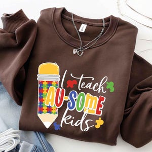 May include: A brown sweatshirt with the text "I teach AU-SOME kids" in colorful letters and a pencil graphic with a puzzle piece design. The sweatshirt has a crew neck and long sleeves. A silver necklace is draped on the sweatshirt.