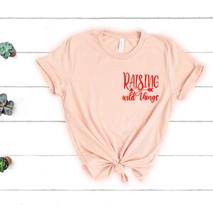 May include: A light pink t-shirt with the text "Raising wild things" in red lettering on the front.