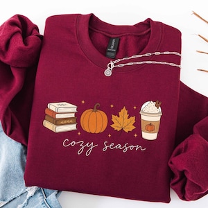 May include: A maroon crewneck sweatshirt featuring a graphic print of a stack of books, an orange pumpkin, a maple leaf, and a coffee cup with whipped cream. The text "cozy season" is printed in white cursive script below the graphic elements.