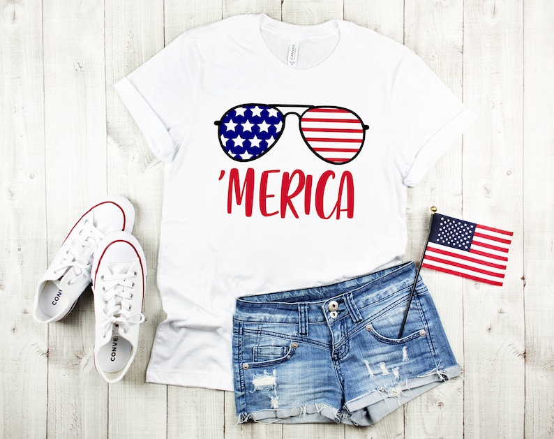 May include: White t-shirt featuring a red, white and blue graphic of sunglasses with the text 'MERICA' on the front. The shirt is paired with denim shorts and white trainers.