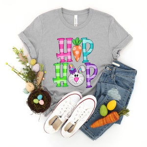 May include: A gray t-shirt with the words "HOP HOP" in colorful block letters, featuring a carrot and a bunny face. The shirt is paired with blue jeans, white sneakers, and Easter decorations, creating a festive spring theme.