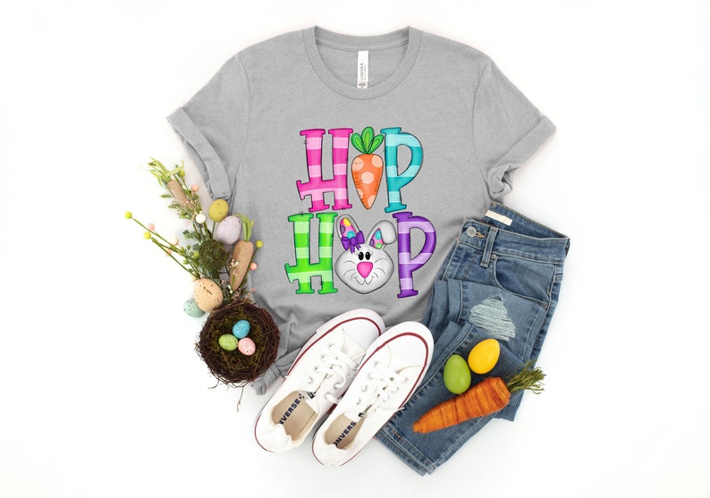 May include: A gray t-shirt with the words "HOP HOP" in colorful block letters, featuring a carrot and a bunny face. The shirt is paired with blue jeans, white sneakers, and Easter decorations, creating a festive spring theme.