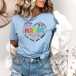 Puede incluir: Una camiseta azul claro con un dise&ntilde;o de coraz&oacute;n colorido que dice "MADRE" rodeado de palabras en espa&ntilde;ol que describen a una madre. Las palabras son "inteligente", "sabia", "cari&ntilde;osa", "fuerte", "buena", "bella" y "valiente".