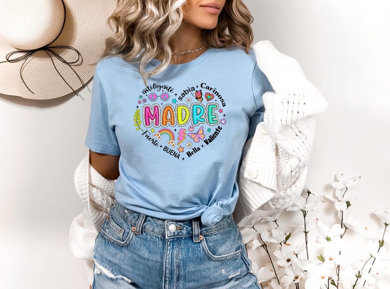 Puede incluir: Una camiseta azul claro con un dise&ntilde;o de coraz&oacute;n colorido que dice "MADRE" rodeado de palabras en espa&ntilde;ol que describen a una madre. Las palabras son "inteligente", "sabia", "cari&ntilde;osa", "fuerte", "buena", "bella" y "valiente".