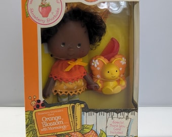 Może przedstawiać: Lalka Strawberry Shortcake Orange Blossom w pomarańczowo-żółtej sukience, żółtym motylem i grzebieniem. Na pudełku widnieje napis "Orange Blossom with Marmalade" i "Doll and Pet Orange Scented Non-Toxic".