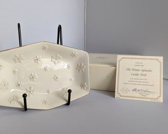May include: A white Lenox 'The Winter Splendor Candy Dish' with a snowflake design. The dish is on a black stand and comes with a certificate of authenticity.