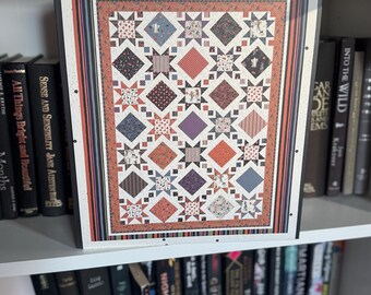 May include: A framed quilt with a patchwork design featuring diamonds and stars in various colors like red, blue, and purple. The quilt is framed in a white border with a striped pattern. The image is displayed on a white shelf with books.