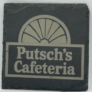 May include: Square, dark gray slate coaster with the words "Putsch's Cafeteria" in a light gray font. Above the text is a light gray semi-circle design. The coaster has a rough, textured surface.