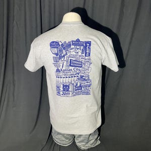May include: A gray t-shirt with a blue graphic featuring various Kansas City landmarks and businesses, including "Italian Gardens," "Wish Bone," and "Katz." The shirt is displayed on a mannequin.