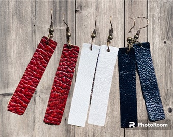 May include: Three pairs of leather earrings in red, white, and black. Each pair features a rectangular leather piece with a textured surface.