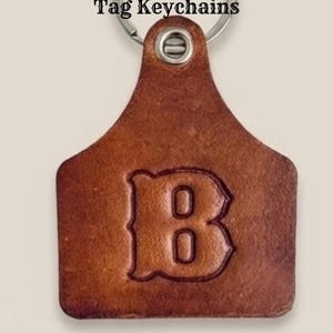 May include: A brown leather keychain shaped like a livestock ear tag with a silver metal ring at the top. The letter B is embossed in the center of the leather. The text Tag Keychains is printed in black above the leather tag.