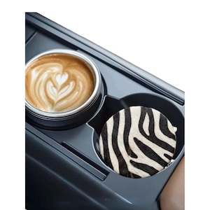May include: A circular car cup holder coaster featuring a black and white zebra stripe pattern. The coaster is placed inside a dark gray plastic vehicle console cup holder next to a silver metal cup filled with a latte featuring heart-shaped foam art.