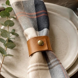 May include: A tan leather napkin ring featuring a centered brass-toned metal snap button closure. The ring secures a folded fabric napkin patterned with a plaid design in shades of cream, charcoal gray, and thin rust-colored accent lines.