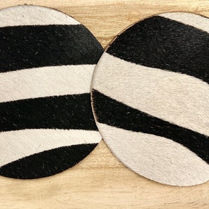 May include: Two round black and white zebra print coasters. The coasters are made of cowhide and have a natural, rustic look.