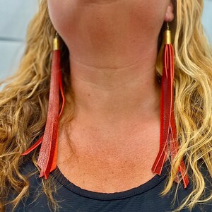May include: A pair of long, red leather tassel earrings. The earrings are hanging from the wearer's ears and are about 10 inches long.