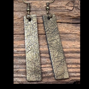 May include: A pair of gold-toned leather earrings with a textured surface. The earrings are rectangular in shape and have a simple hook closure.