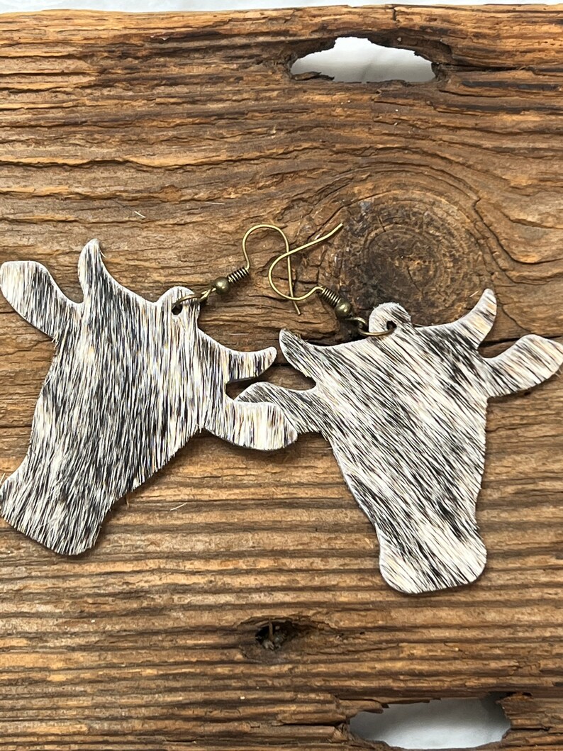 Puede incluir: Un par de pendientes de piel de vaca con forma de cabeza de vaca. Los pendientes son negros y blancos con un gancho de metal marr&oacute;n.