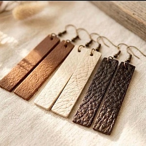 May include: Three pairs of rectangular drop earrings featuring textured surfaces in shades of light brown, cream, and dark brown. Each earring is suspended from a simple metal hook, showcasing a long, slender, flat design with visible grain patterns.