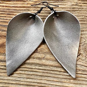 May include: A pair of silver leather earrings shaped like teardrops. The earrings are attached to black metal hooks.