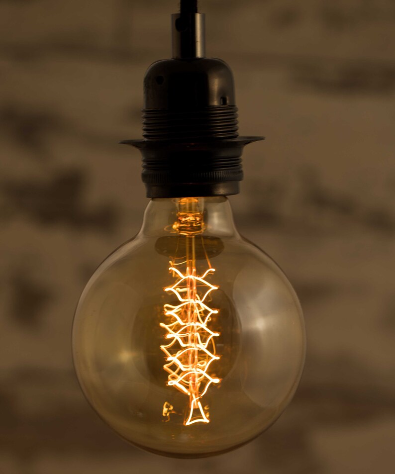 May include: A clear glass light bulb with a decorative filament in the shape of a spiral. The bulb is hanging from a black metal fixture.