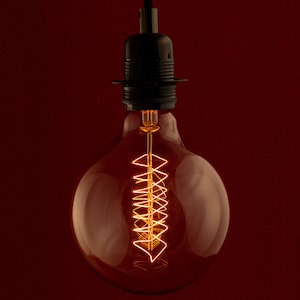 May include: A clear glass light bulb with a coiled filament, lit and glowing with a warm orange light. The bulb is attached to a black metal socket.