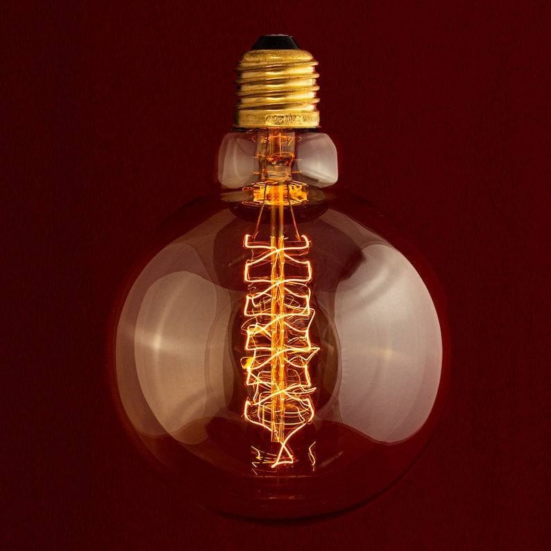 May include: A clear glass light bulb with a gold metal base and a coiled filament inside. The bulb is lit and glowing orange.
