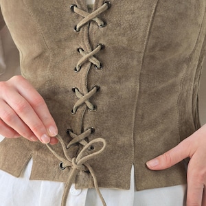May include: A taupe suede vest featuring a front lace-up closure with dark metal grommets and matching suede cord ties. The vest is worn over a white collared shirt with visible buttons, showcasing a structured fit with vertical seam detailing.