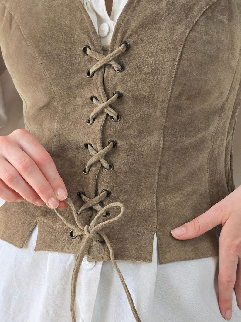 May include: A taupe suede vest featuring a front lace-up closure with dark metal grommets and matching suede cord ties. The vest is worn over a white collared shirt with visible buttons, showcasing a structured fit with vertical seam detailing.