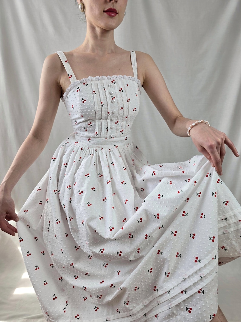 May include: A white midi dress with a cherry print and a lace trim. The dress has a fitted bodice and a full skirt.