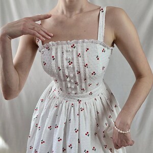 May include: A white sundress with a cherry print and a fitted bodice. The dress has a square neckline and thin straps.