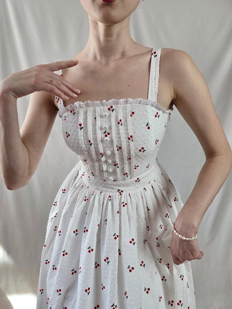 May include: A white sundress with a cherry print and a fitted bodice. The dress has a square neckline and thin straps.