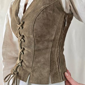 May include: A sleeveless olive brown suede vest featuring a front lace-up closure with matching suede ties, metal grommets, and a side zipper. The garment is constructed with vertical paneling and a straight hemline with small side slits.